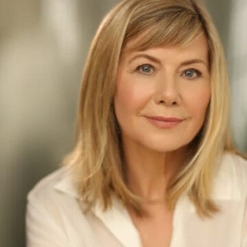 A middle-aged woman with straight blonde hair and a white blouse, resembling Glynis Barber, is looking at the camera with a neutral expression against a blurred background.