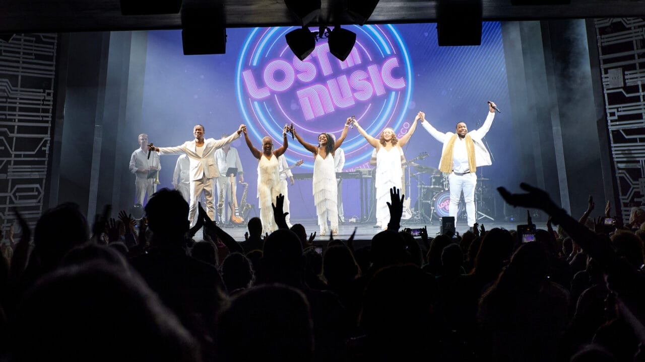 A band dressed in white stands on stage holding hands, facing a cheering audience, with a neon "Lost in Music" sign glowing behind them—a perfect scene from One Night at the Disco.
