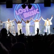 A band dressed in white stands on stage holding hands, facing a cheering audience, with a neon "Lost in Music" sign glowing behind them—a perfect scene from One Night at the Disco.