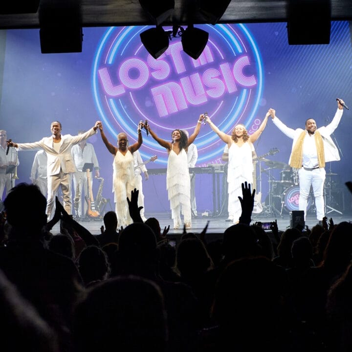 A band dressed in white stands on stage holding hands, facing a cheering audience, with a neon "Lost in Music" sign glowing behind them—a perfect scene from One Night at the Disco.
