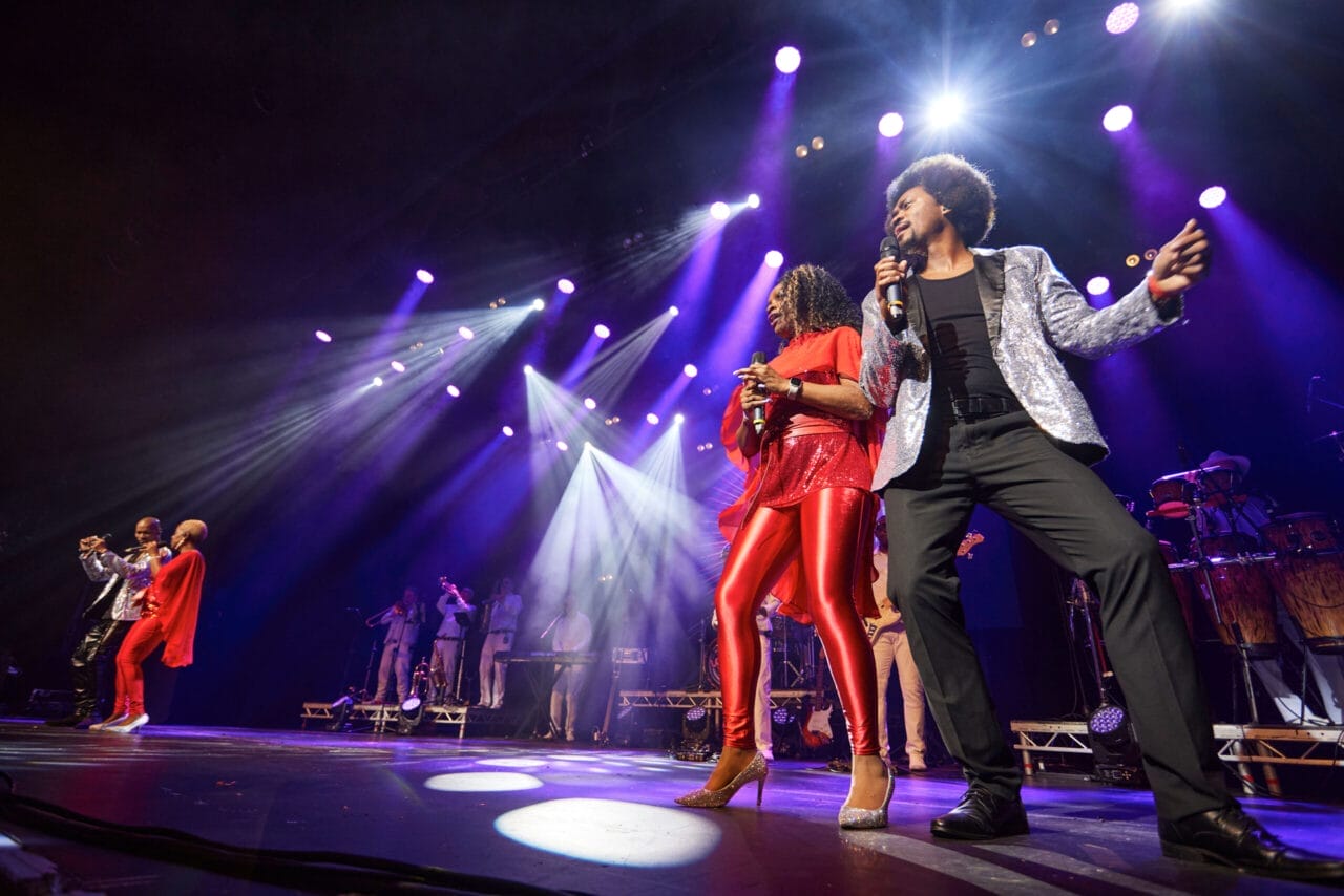 A group of musicians and singers perform energetically on a brightly lit stage, transporting the crowd back to a disco era—it's truly One Night at the Disco with spotlights and groovy instruments.