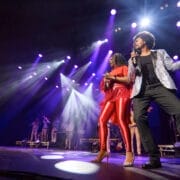 A group of musicians and singers perform energetically on a brightly lit stage, transporting the crowd back to a disco era—it's truly One Night at the Disco with spotlights and groovy instruments.