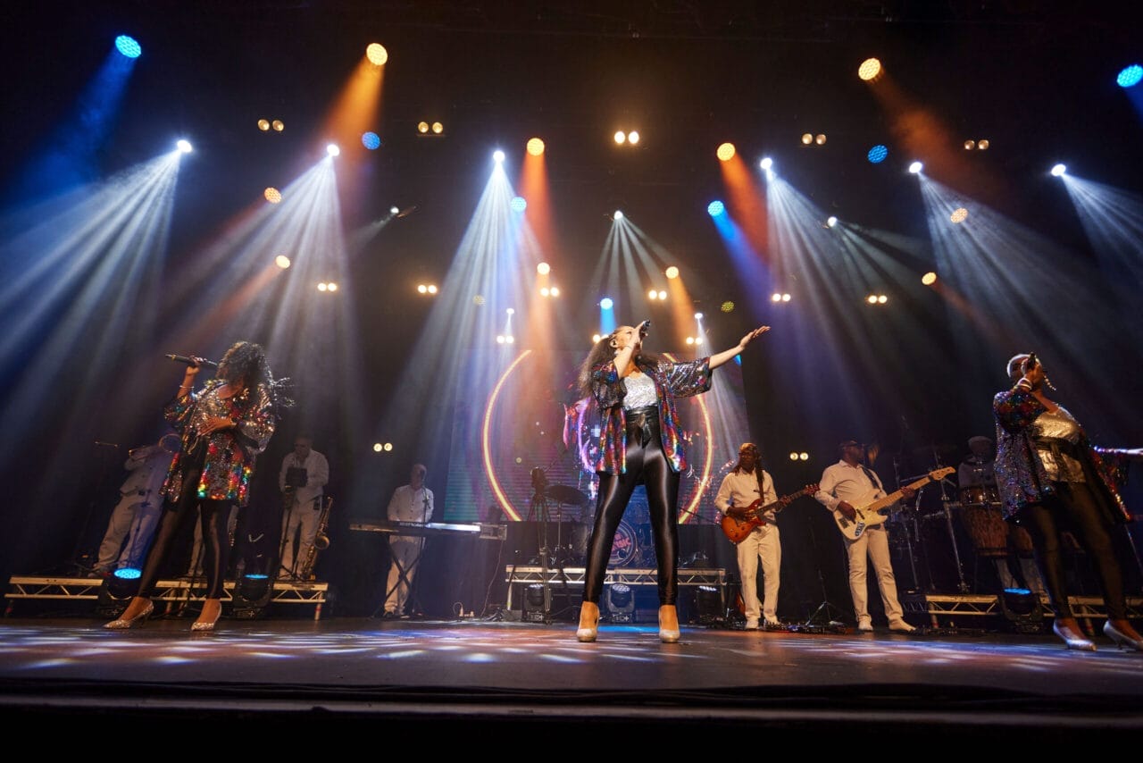 A band performs on stage under colorful lights, with singers in sparkly outfits and musicians playing instruments in the background, creating a true One Night at the Disco experience.