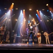 A band performs on stage under colorful lights, with singers in sparkly outfits and musicians playing instruments in the background, creating a true One Night at the Disco experience.