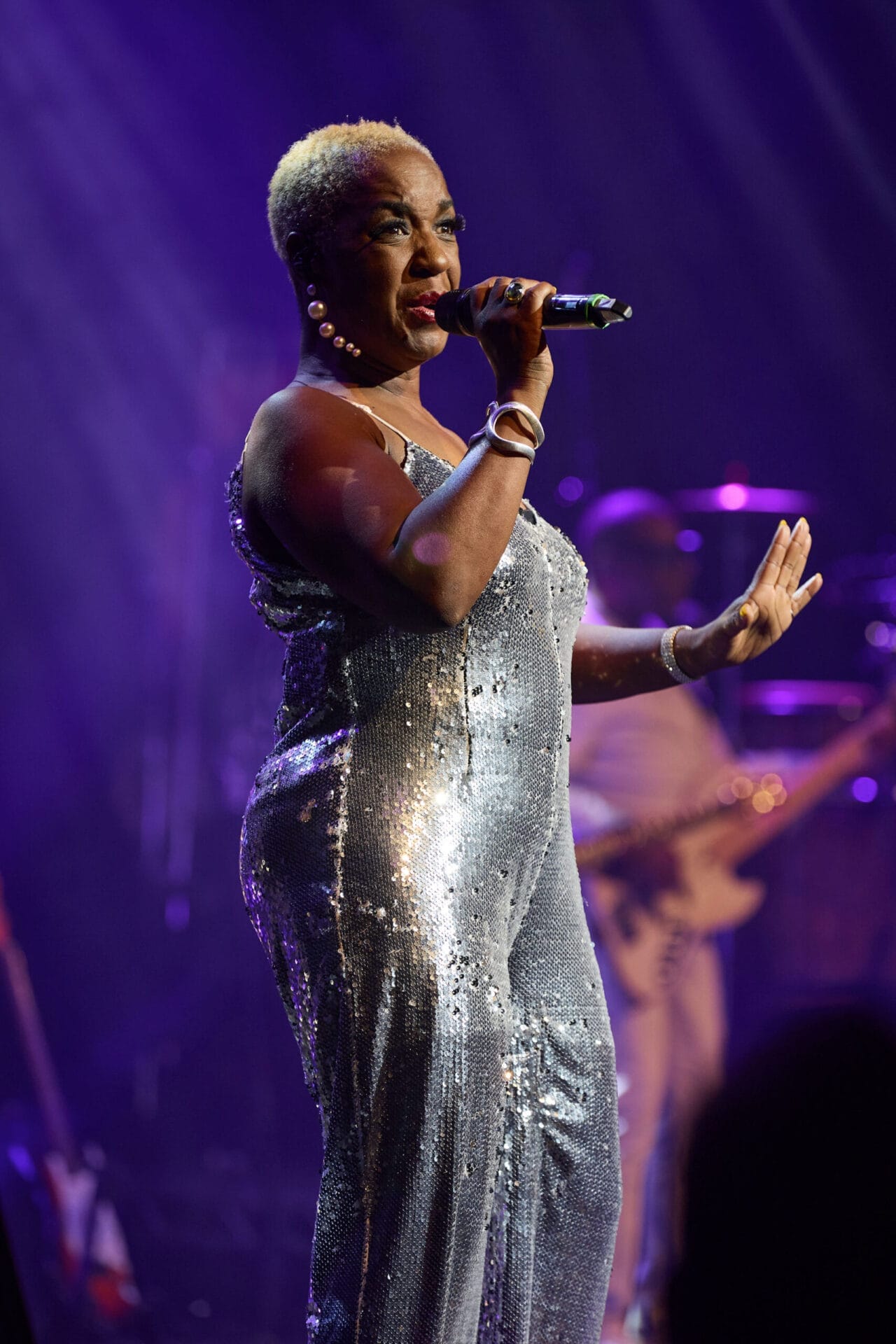 A woman in a silver sequin dress sings into a microphone on stage under purple lighting, capturing the essence of Disco as she performs “Lost in Music” at One Night at the Disco.