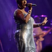 A woman in a silver sequin dress sings into a microphone on stage under purple lighting, capturing the essence of Disco as she performs “Lost in Music” at One Night at the Disco.