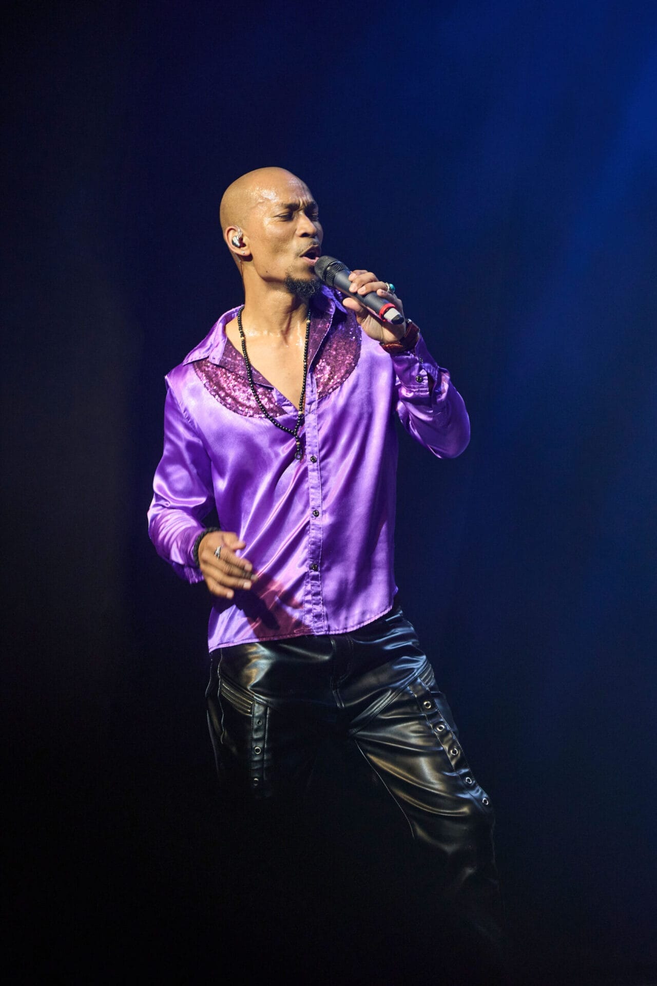 A man in a shiny purple shirt and black pants sings into a microphone on stage under blue lighting, looking lost in music as if it's one night at the disco.