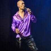 A man in a shiny purple shirt and black pants sings into a microphone on stage under blue lighting, looking lost in music as if it's one night at the disco.
