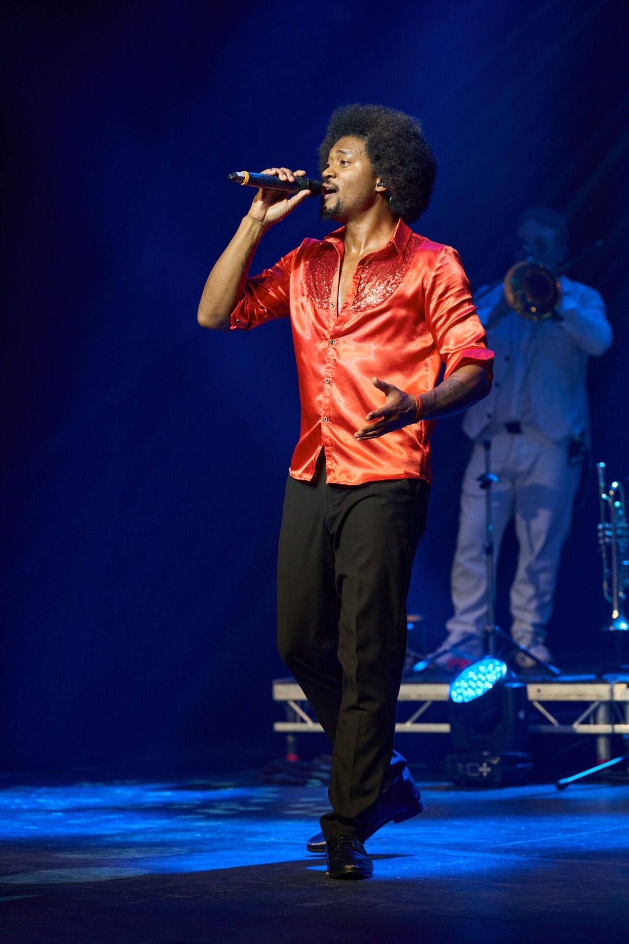 A man in a bright red shirt sings into a microphone on stage during One Night at the Disco, while a musician in white plays the trombone in the background.