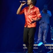 A man in a bright red shirt sings into a microphone on stage during One Night at the Disco, while a musician in white plays the trombone in the background.