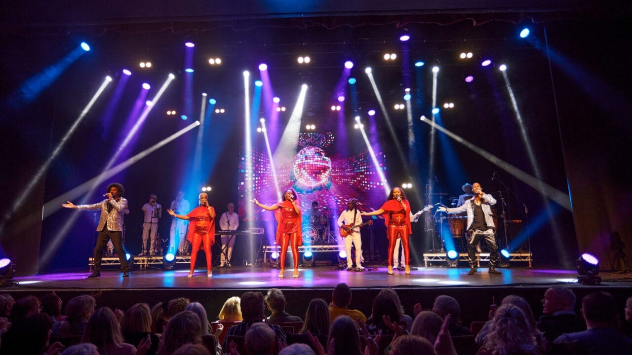 A music group performs on stage under colorful lights, captivating a large audience seated in front; a screen with visual effects sets the mood for a night lost in music, evoking the spirit of One Night at the Disco.