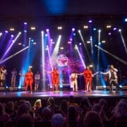 A music group performs on stage under colorful lights, captivating a large audience seated in front; a screen with visual effects sets the mood for a night lost in music, evoking the spirit of One Night at the Disco.