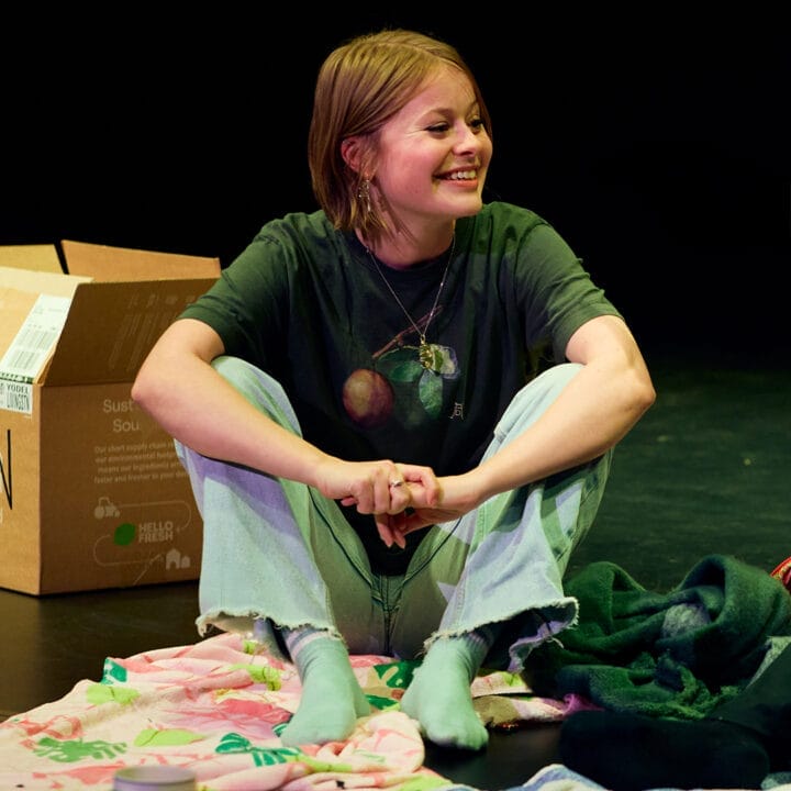 A young person sits cross-legged on the floor, smiling, with a cardboard box and colorful blankets nearby.