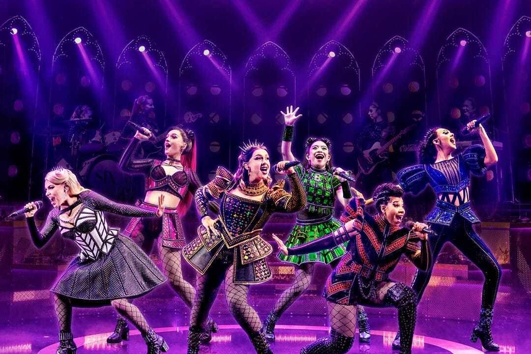 Six performers in colorful, stylized costumes sing and pose energetically on stage under purple lighting, with a live band in the background.
