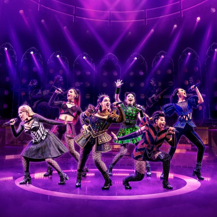 Six performers in colorful, stylized costumes sing and pose energetically on stage under purple lighting, with a live band in the background.