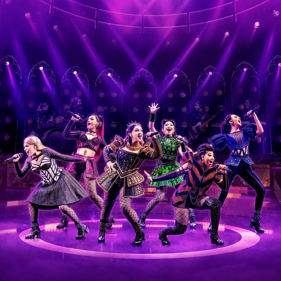 Six performers in colorful, stylized costumes sing and pose energetically on stage under purple lighting, with a live band in the background.