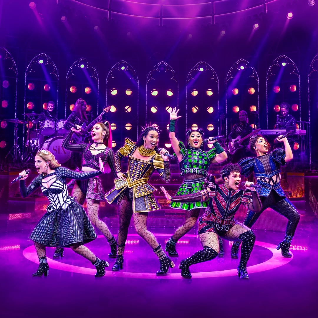 Six performers in colorful, stylized costumes sing and pose energetically on stage under purple lighting, with a live band in the background.