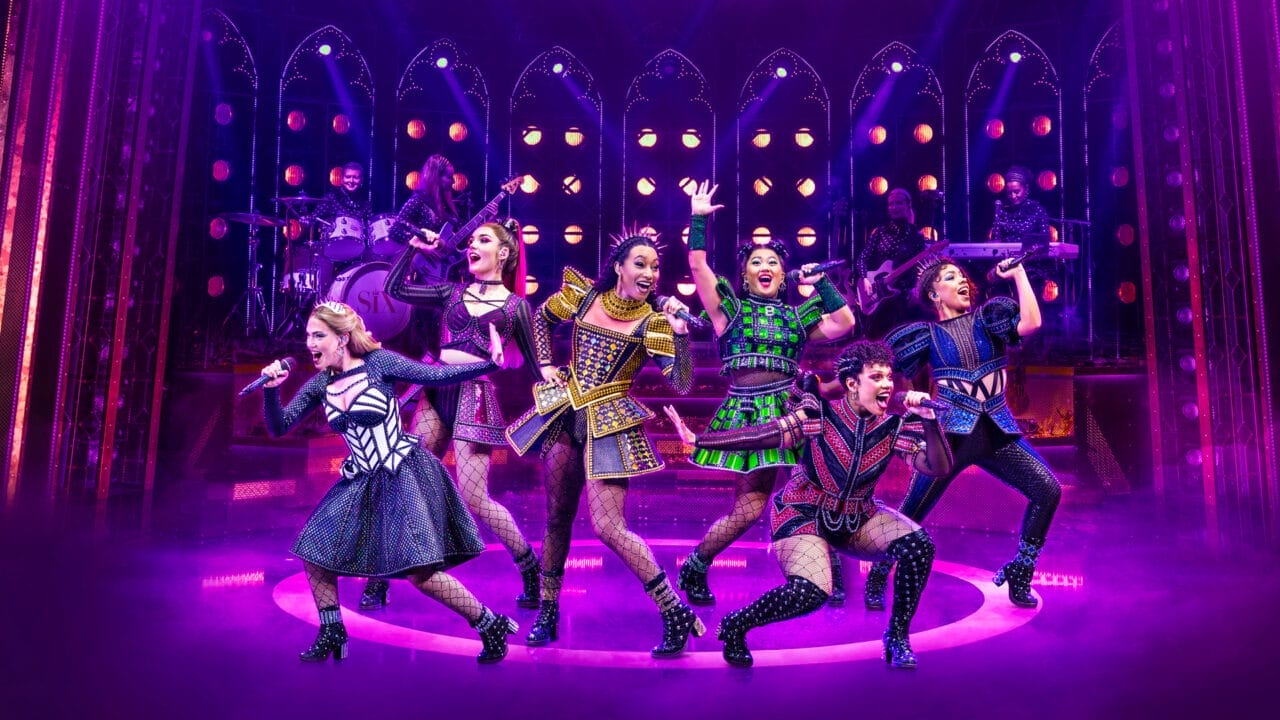Six performers in colorful, stylized costumes sing and pose energetically on stage under purple lighting, with a live band in the background.