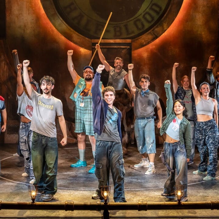 A group of actors in casual costumes stand on stage with one arm raised.
