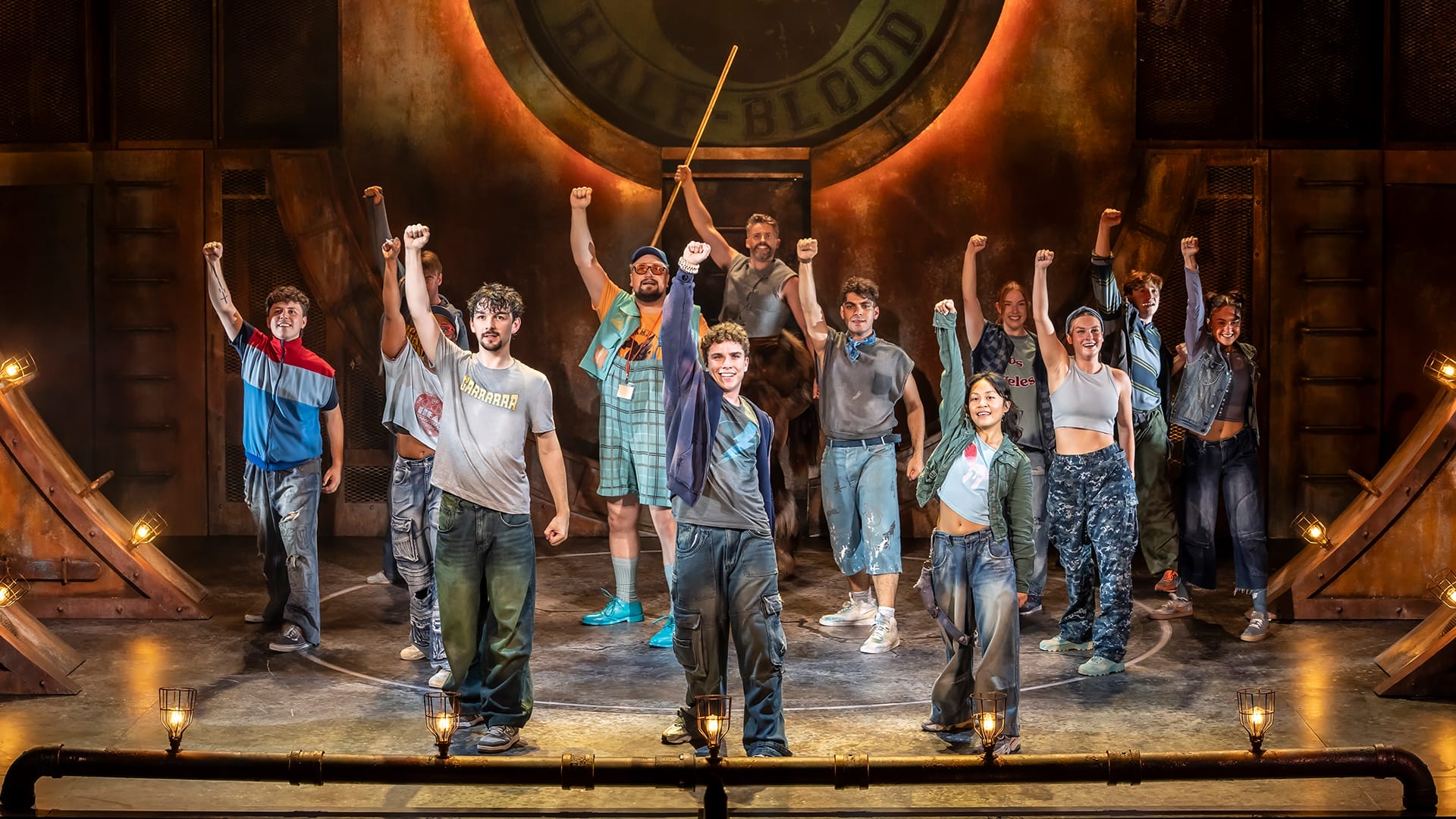 A group of actors in casual costumes stand on stage with one arm raised.