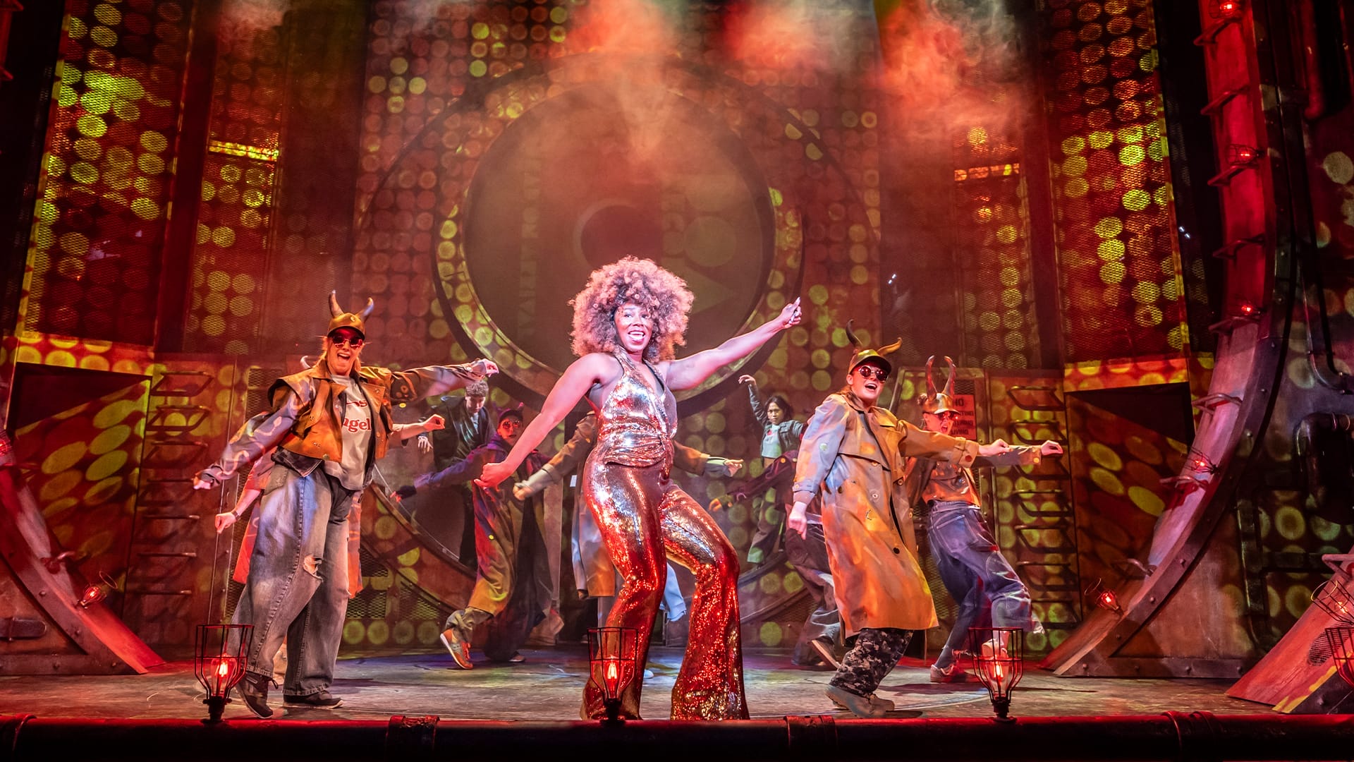 A performer in a shiny gold jumpsuit sings and dances center stage, showcasing classic Musical Theatre flair with backup dancers in trench coats and hats, under dramatic stage lighting and a bold circular backdrop.
