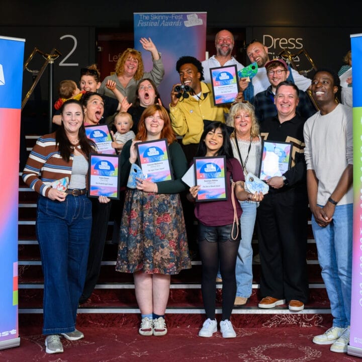 A group of people smile and hold certificates and trophies on a staircase between two "The Skinny-Fest Festival Awards" banners, celebrating the 2025 Besties awards as some of the first winners at this indoor event.