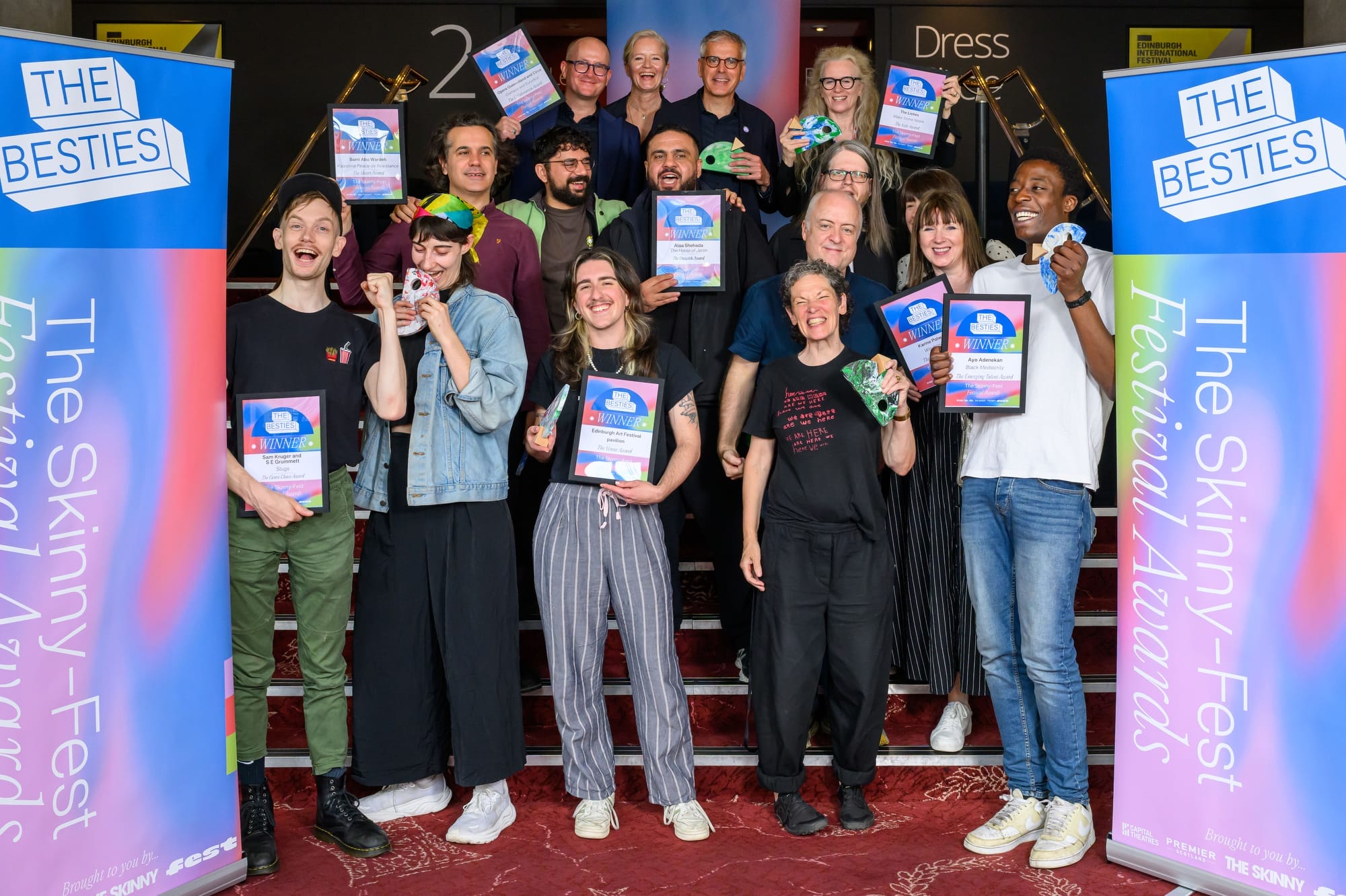A group of people stand on stairs holding certificates and trophies, smiling for a photo at The Skinny-Fest Festival Awards, celebrating as 2025 Besties winners with "The Besties" banners on each side.