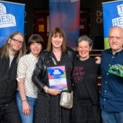 Five people stand together indoors at an awards event, with one person holding a winner’s certificate and another displaying a green, leaf-shaped trophy as the week two winners of the 2025 Besties.
