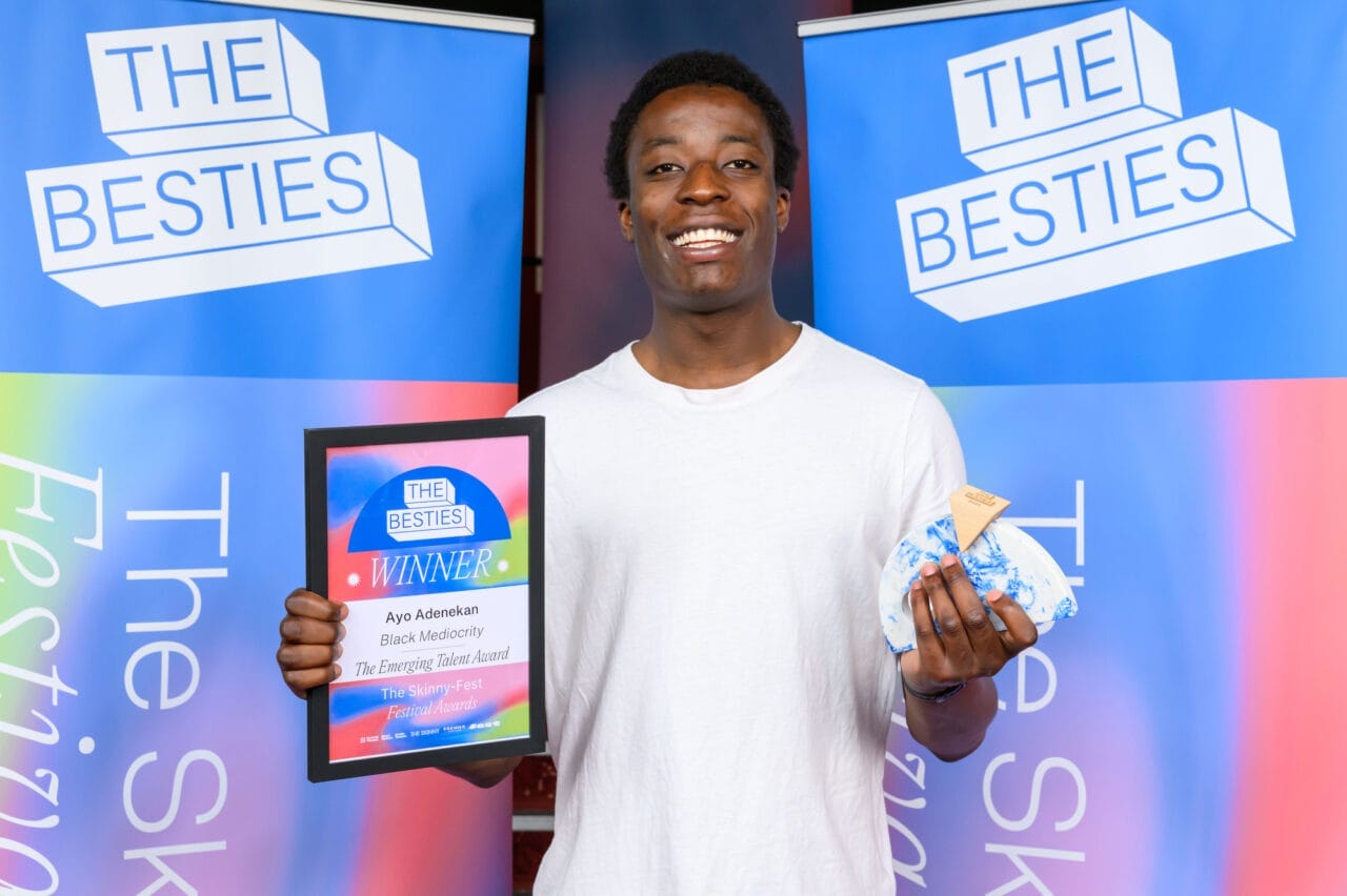 A person in a white shirt holds a winner’s plaque and a wrapped item, standing in front of banners that read “THE BESTIES,” celebrating the 2025 Besties week two winners.