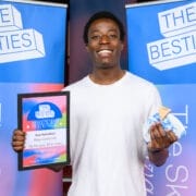 A person in a white shirt holds a winner’s plaque and a wrapped item, standing in front of banners that read “THE BESTIES,” celebrating the 2025 Besties week two winners.