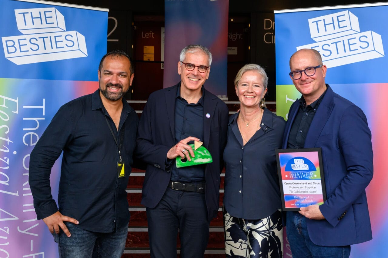 Four people stand smiling, holding an award and a certificate, in front of banners reading "The Besties" at the 2025 Besties event, celebrating as week two winners at the festival awards ceremony.