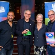 Four people stand smiling, holding an award and a certificate, in front of banners reading "The Besties" at the 2025 Besties event, celebrating as week two winners at the festival awards ceremony.
