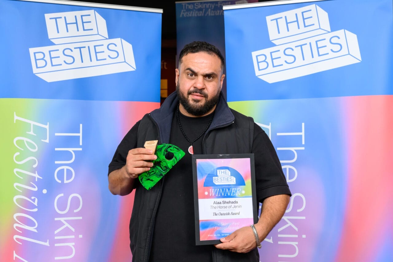 A man holds a framed certificate and a small green item, standing in front of blue banners reading "The Besties" at an awards event celebrating Besties winners.
