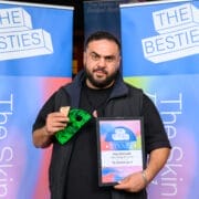 A man holds a framed certificate and a small green item, standing in front of blue banners reading "The Besties" at an awards event celebrating Besties winners.