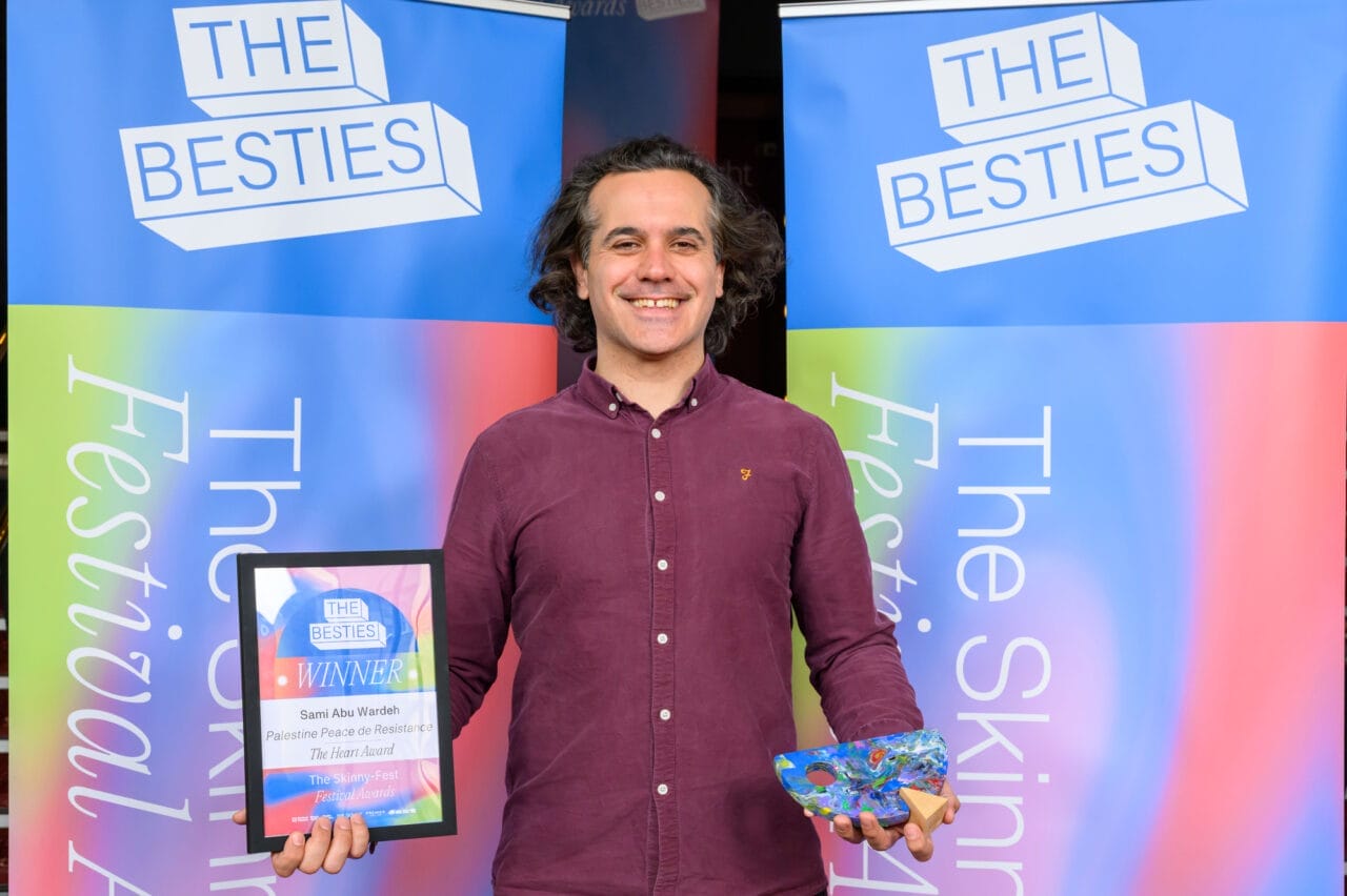A person stands smiling, holding a winner's plaque and a colorful box in front of two "The Besties" festival award banners, celebrating as one of the 2025 Besties week two winners.