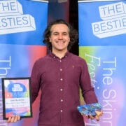 A person stands smiling, holding a winner's plaque and a colorful box in front of two "The Besties" festival award banners, celebrating as one of the 2025 Besties week two winners.