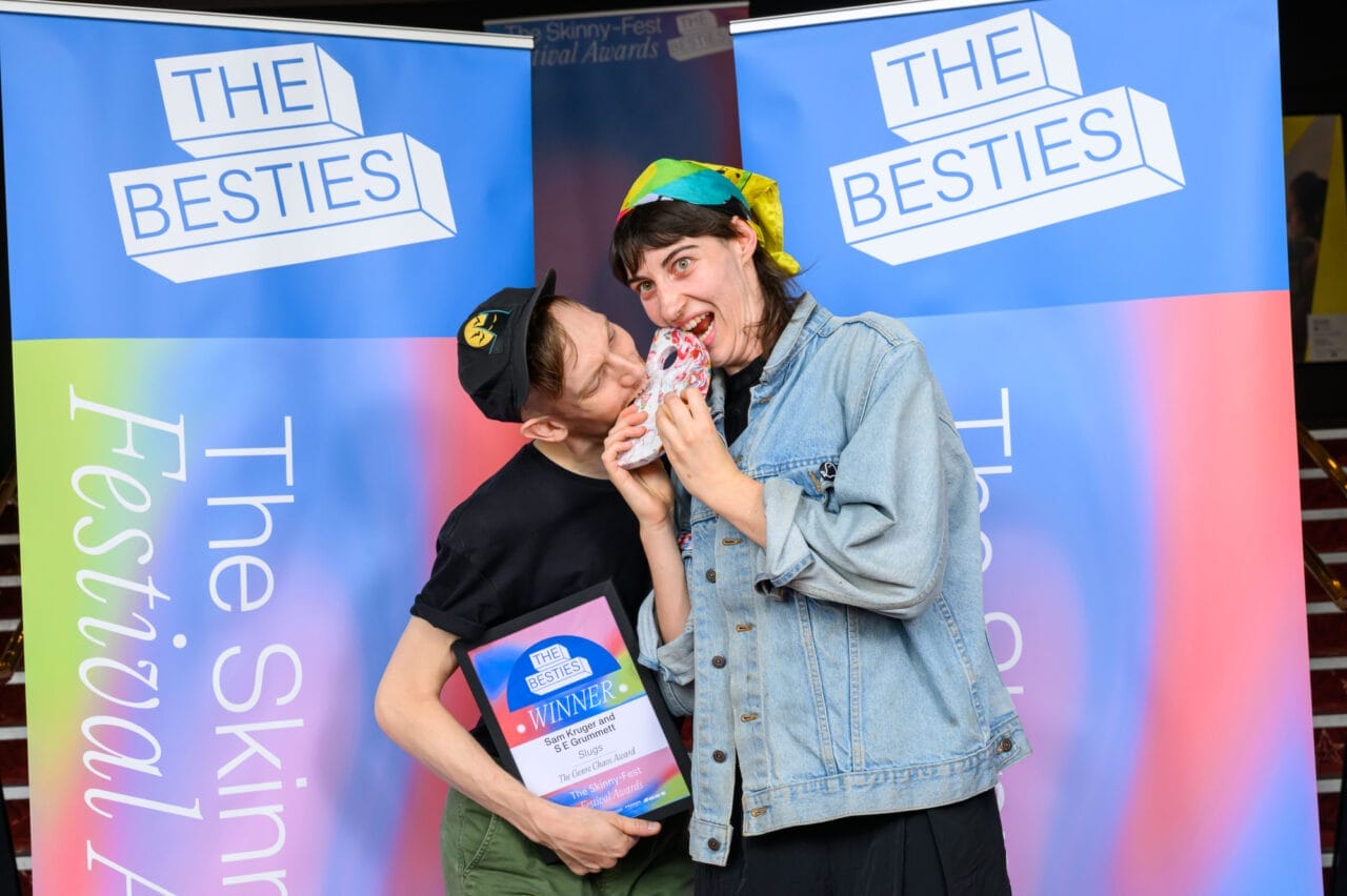 Two people pose in front of "The Besties" banners; one, a 2025 Besties week two winner, playfully bites the other's shoulder while holding a plaque, as the other smiles with a patterned item.