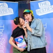 Two people pose in front of "The Besties" banners; one, a 2025 Besties week two winner, playfully bites the other's shoulder while holding a plaque, as the other smiles with a patterned item.