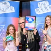 Three people smile and pose in front of "The Besties" banners; the center person holds a 2025 Besties winner's plaque while the others display week two winners lanyards and a colorful packet.