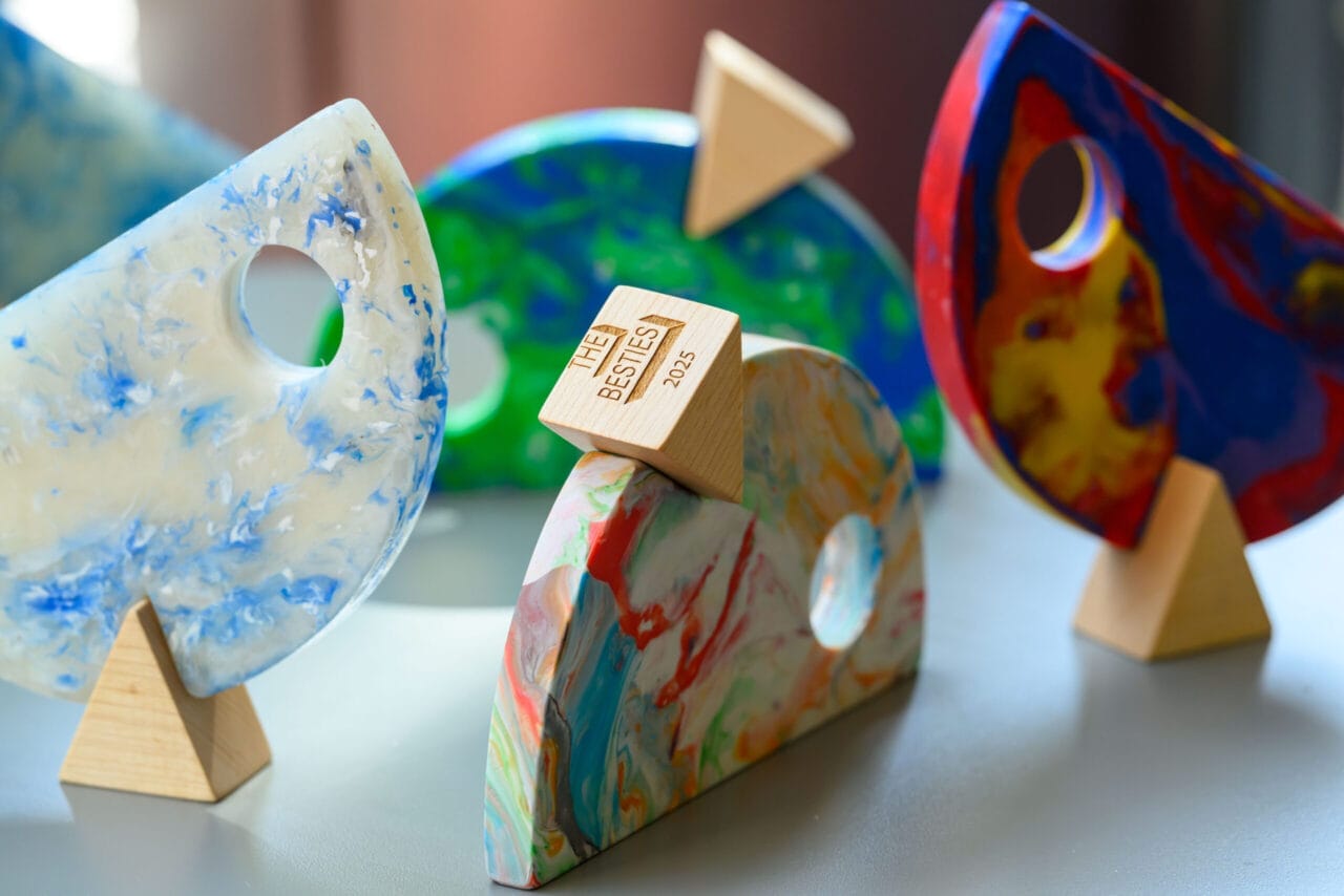 Four colorful, abstract sculptural awards with circular cutouts and wooden bases are displayed on a table; one bears a small plaque reading "THE BESTIES 2023," celebrating the first winners ahead of the 2025 Besties.