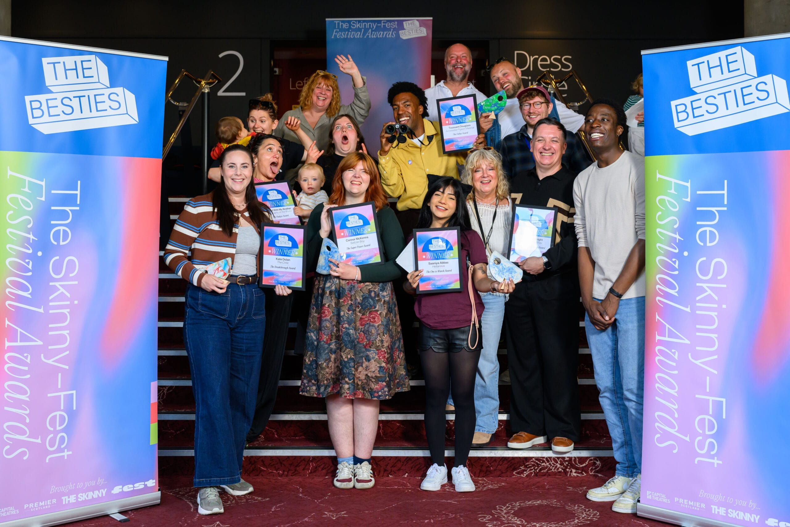 A group of people smile and hold certificates and trophies on a staircase between two "The Skinny-Fest Festival Awards" banners, celebrating the 2025 Besties awards as some of the first winners at this indoor event.