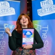 A person smiling and holding a glass award and a framed certificate in front of two banners that read "THE BESTIES," celebrating as one of the first winners of the 2025 Besties.