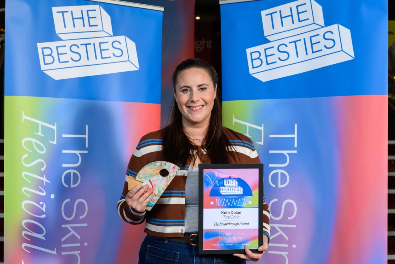 A woman stands in front of "The Besties" banners, holding an award plaque and a small trophy, celebrating as one of the first winners of the 2025 Besties.