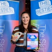 A woman stands in front of "The Besties" banners, holding an award plaque and a small trophy, celebrating as one of the first winners of the 2025 Besties.