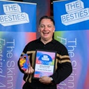 A man stands smiling, holding a colorful award and a framed certificate, in front of banners that read "2025 Besties" and "The Skinny Festival Awards," celebrating the Besties winners.