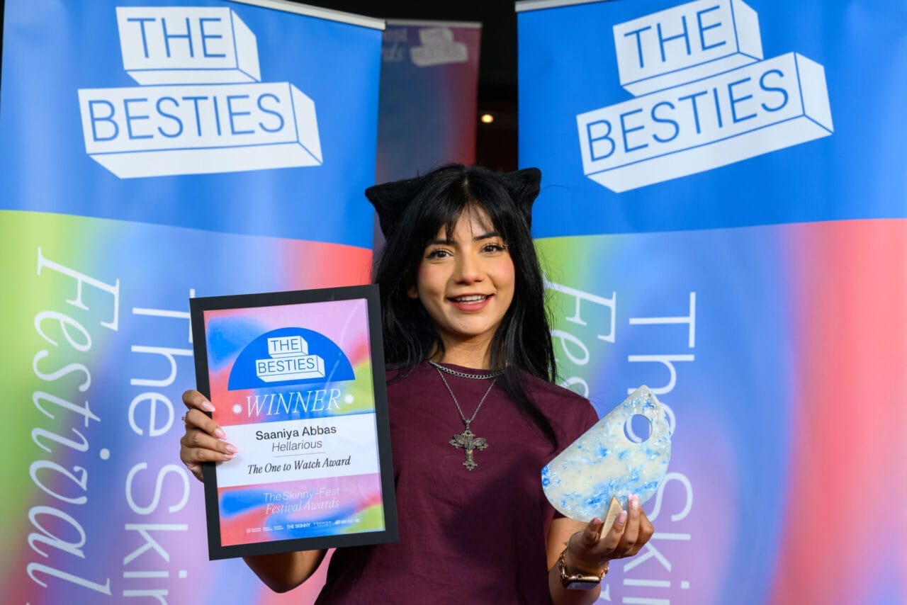 A woman holding an award plaque and trophy stands in front of banners that read "The Besties" and "The Skinny Fest," celebrating with fellow 2025 Besties winners.