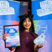 A woman holding an award plaque and trophy stands in front of banners that read "The Besties" and "The Skinny Fest," celebrating with fellow 2025 Besties winners.