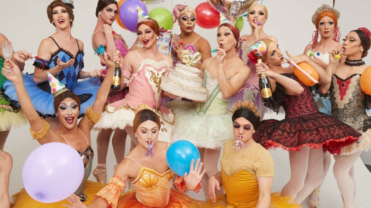 A group inspired by Les Ballets Trockadero de Monte Carlo, dressed in colorful ballet costumes and party hats, hold balloons, cake, and party favors as they celebrate together in a lively and festive setting.