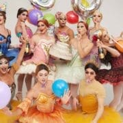 A group inspired by Les Ballets Trockadero de Monte Carlo, dressed in colorful ballet costumes and party hats, hold balloons, cake, and party favors as they celebrate together in a lively and festive setting.
