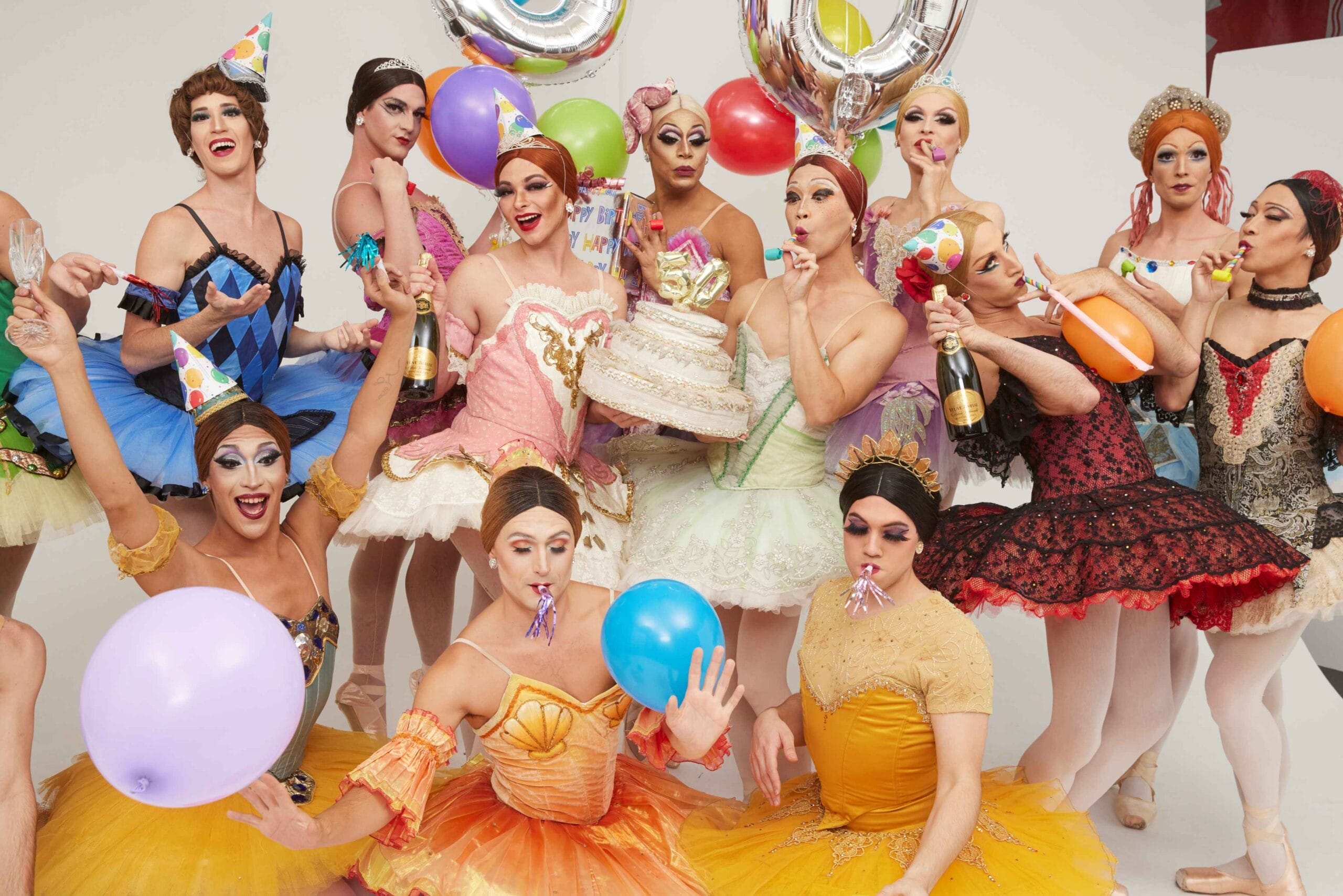 A group inspired by Les Ballets Trockadero de Monte Carlo, dressed in colorful ballet costumes and party hats, hold balloons, cake, and party favors as they celebrate together in a lively and festive setting.